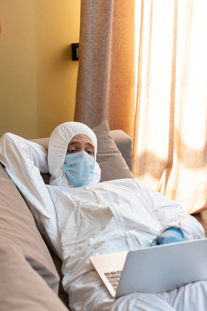 Selective focus of freelancer in medical mask and hazmat suit using laptop while lying on couch at home  - Photo, Image