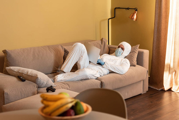 KYIV, UKRAINE - APRIL 24, 2020: Selective focus of man in hazmat suit and medical mask using laptop near joystick and remote controller on couch  - Fotó, kép