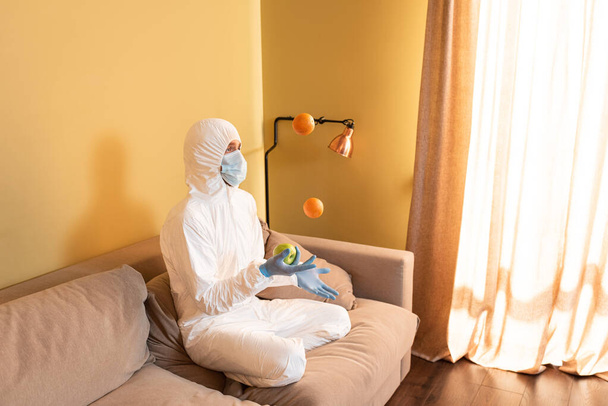 Mann in Warnanzug und Schutzmaske jongliert mit Früchten auf Couch  - Foto, Bild