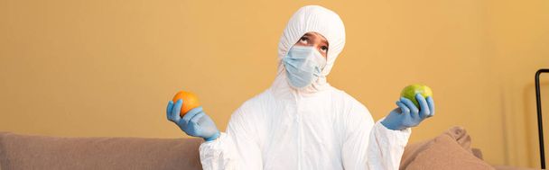 Panoramic orientation of thoughtful man in hazmat suit and medical mask holding apple and orange at home  - Photo, Image