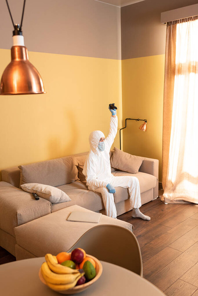 KYIV, UKRAINE - APRIL 24, 2020: Selective focus of man in hazmat suit and medical mask holding joystick on sofa at home  - Foto, immagini