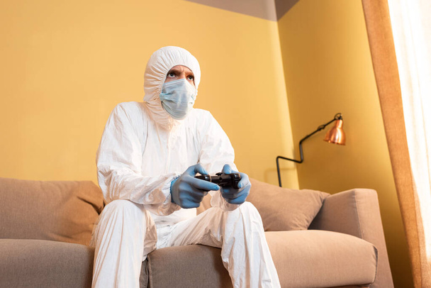 KYIV, UKRAINE - APRIL 24, 2020: Low angle view of man in medical mask and hazmat suit playing video game at home  - Фото, изображение
