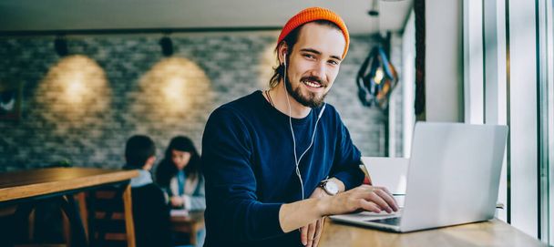 Portrait of happy smiling hipster guy enjoying time for favourite music playlist during e learning in cafeteria, positive male teenager in headphones rest indoors sitting at table with laptop device - Φωτογραφία, εικόνα
