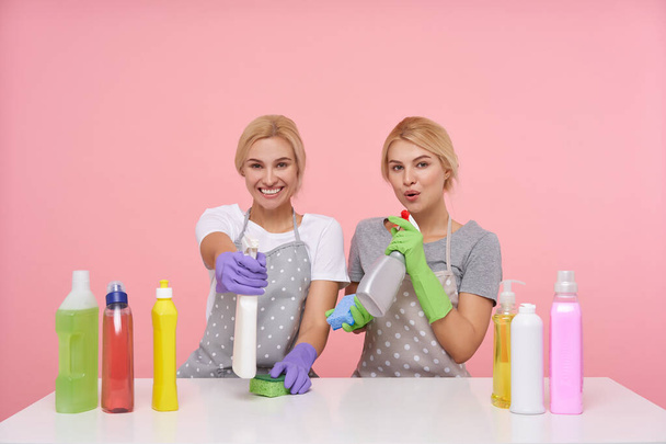 Indoor photo of lovely young white-headed housewives being in nice mood while making spring cleaning, sitting over pink background with household chemicals - Photo, Image
