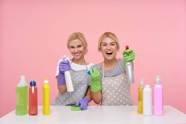 Szczęśliwa młoda atrakcyjna blondynka sprzątająca panie ubrane w mundur podnosząc ręce z butelkami sprayu i uśmiechając się chętnie przed kamerą, izolowane na różowym tle - Zdjęcie, obraz