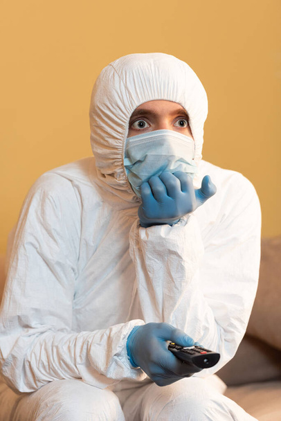 Egy férfi védőruhában, orvosi maszkban és gumikesztyűben távirányítót tart a kanapén.  - Fotó, kép