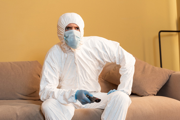 Удумливий чоловік у костюмі та медичній масці дивиться телевізор на дивані
  - Фото, зображення