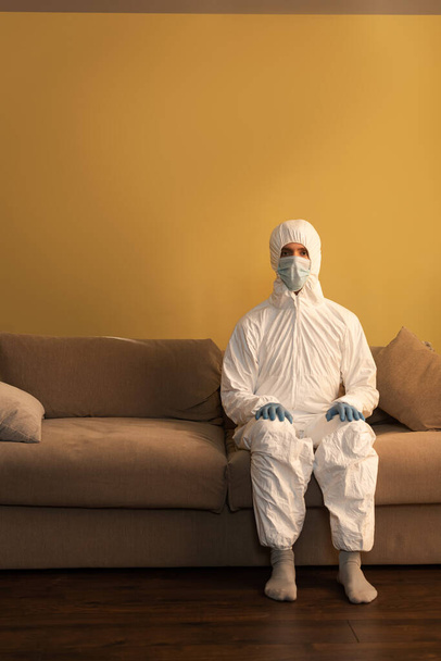 Hombre con traje de materiales peligrosos, máscara médica y guantes de látex mirando a la cámara mientras está sentado en el sofá
  - Foto, Imagen