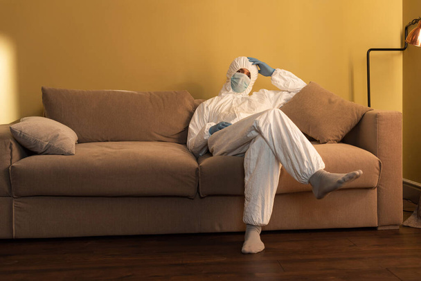 Άντρας με ιατρική μάσκα, στολή απολύμανσης και γάντια λατέξ που κάθονται στον καναπέ στο σπίτι  - Φωτογραφία, εικόνα