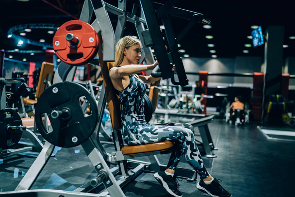 Side view of attractive blonde fit female bodybuilder in gray camouflage sportswear  lifting heavy weights with chest machine in modern gym - Φωτογραφία, εικόνα