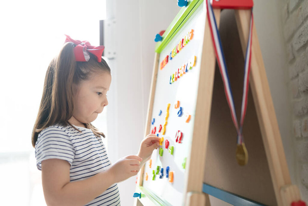 Profiel weergave van een schattig klein meisje met behulp van een magneetbord om letters en cijfers te leren thuis - Foto, afbeelding