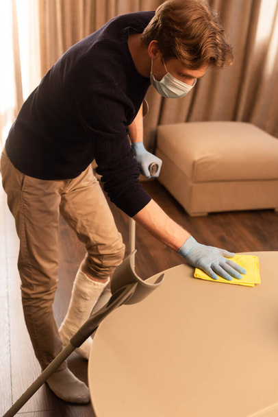 Vista laterale dell'uomo in maschera medica e bendaggio in gesso sul tavolo di pulizia delle gambe in soggiorno
 - Foto, immagini