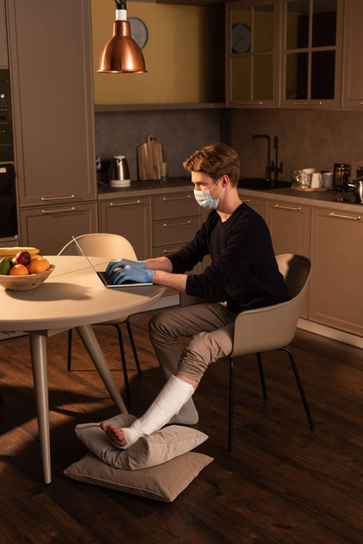 Side view of freelancer in medical mask with broken leg working on laptop in kitchen  - Photo, Image