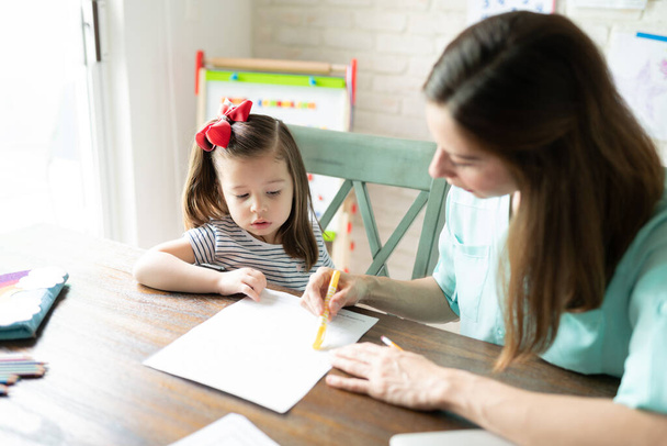 Mutter erklärt eine Aktivität und gibt ihrer kleinen Tochter Anweisungen, während sie unterrichtet und Hausaufgaben macht - Foto, Bild