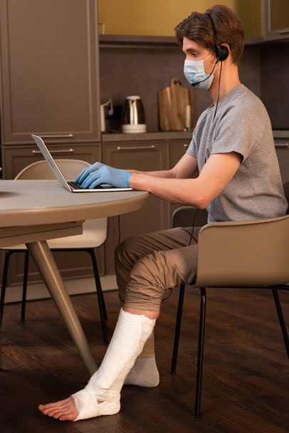 Vista laterale del telelavoratore in maschera medica, guanti in lattice e bendaggio in gesso sulla gamba utilizzando cuffie mentre si lavora su laptop in cucina
  - Foto, immagini