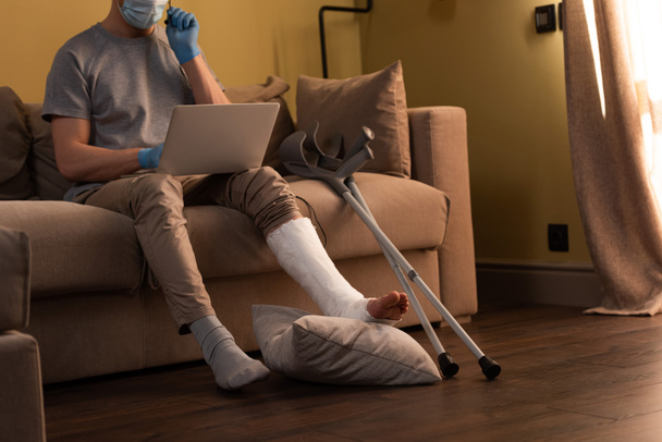 Vágott kilátás férfi törött láb és orvosi maszk segítségével laptop és fejhallgató közelében mankók kanapén  - Fotó, kép