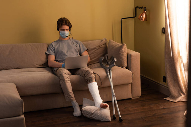 自宅のノートパソコンで作業中にヘッドセットを使用して骨折した脚を持つ医療用マスクの男  - 写真・画像