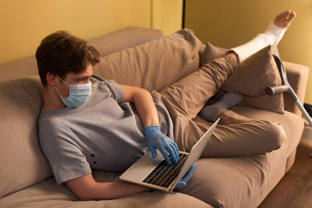 Concentration sélective de l'homme dans le masque médical et le bandage de plâtre sur la jambe à l'aide d'un ordinateur portable sur le canapé
  - Photo, image