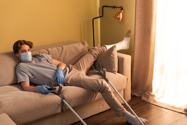 Disabled man in medical mask and plaster bandage on leg watching movie near crutches at home  - Фото, изображение