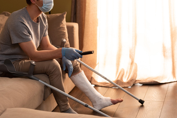 Widok człowieka w masce medycznej i złamanej nogi klikając kanały w domu   - Zdjęcie, obraz