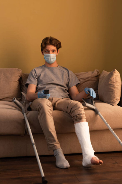 Мужчина в медицинской маске и с гипсовой повязкой на ногах смотрит телевизор, сидя дома на диване
 - Фото, изображение