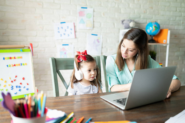 Schattig klein meisje en haar moeder het dragen van een koptelefoon en met behulp van een laptop computer voor een aantal interactieve activiteiten en leren thuis - Foto, afbeelding