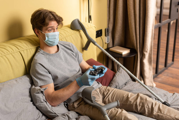 KYIV, UKRAINE - APRIL 24, 2020: Disabled man in medical mask and latex gloves holding gamepad near crutches in bedroom - Foto, afbeelding