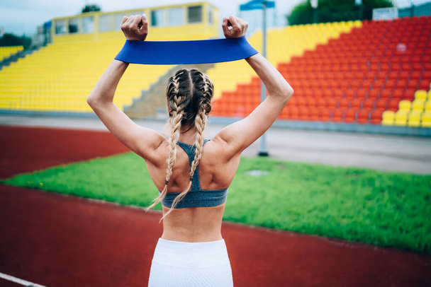 Back view of unrecognizable blond woman standing on stadium with arms above head and stretching blue elastic ribbon during workout - Foto, Bild