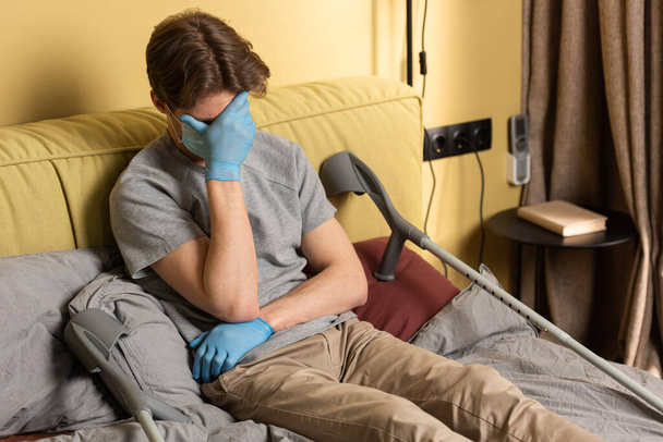 Hombre deprimido en máscara médica y guantes de látex cubriendo la cara con la mano cerca de muletas en la cama
  - Foto, Imagen