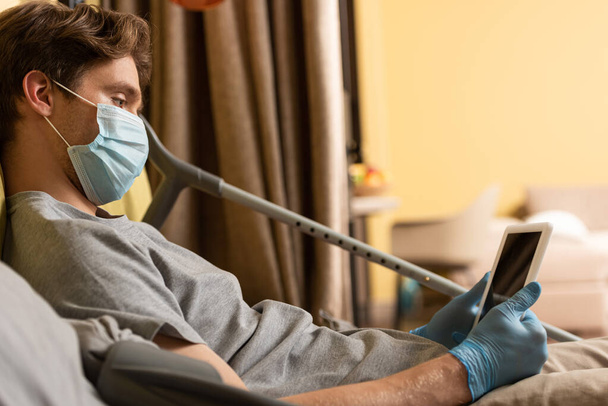 Selective focus of disabled man in medical mask using laptop while lying near crutches on bed  - Photo, Image