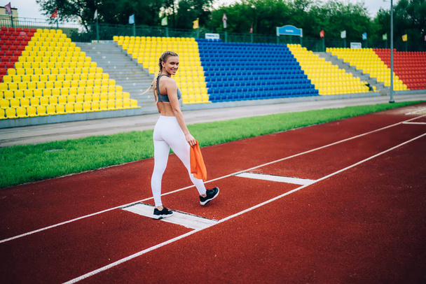 Side view of adult sporty female in modern sportive wear holding rubber resistance band while walking on track of city stadium against colorful grandstand smiling to camera  - 写真・画像