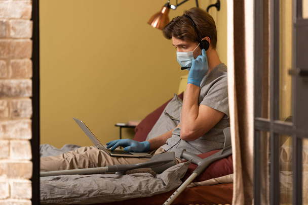 Selective focus of freelancer in medical mask and headset using laptop near crutches on bed  - Photo, Image