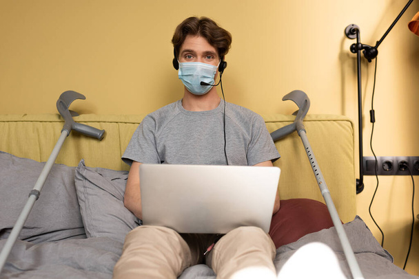 Selective focus of man in medical mask and headset looking at camera while holding laptop near crutches on bed  - Foto, Imagen