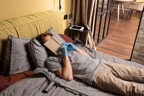 Man in latex gloves covering face with book near crutches on bed  - Fotoğraf, Görsel