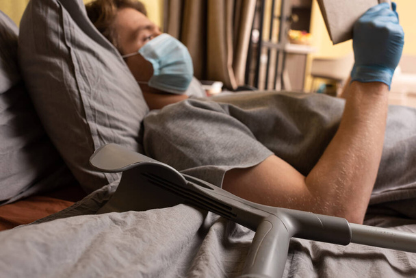 Szelektív fókusz a fogyatékkal élő ember orvosi maszk olvasókönyv közelében mankó az ágyban  - Fotó, kép