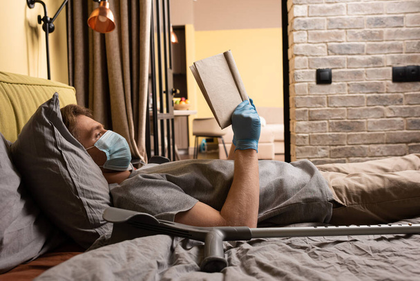 Vista lateral del hombre en máscara médica y guantes de látex libro de lectura cerca de muleta en la cama
  - Foto, Imagen