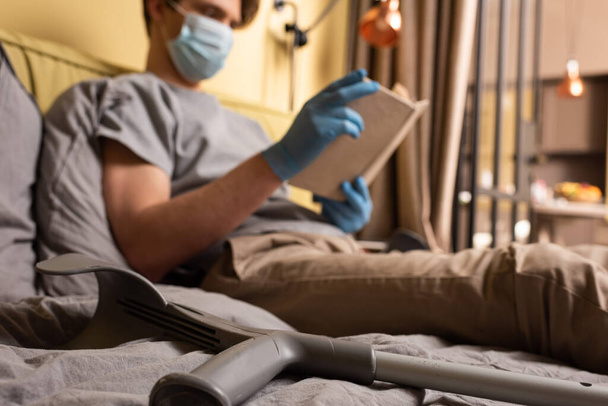 Tıbbi maskeli bir adamın yanında koltuk değneklerinin seçici odağı ve yatakta kitap okuyan lateks eldivenler  - Fotoğraf, Görsel
