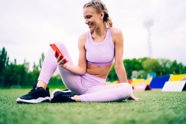 Smiling young sporty woman in modern sportive wear surfing smartphone while sitting on green grass of soccer field of city stadium having rest - Fotoğraf, Görsel