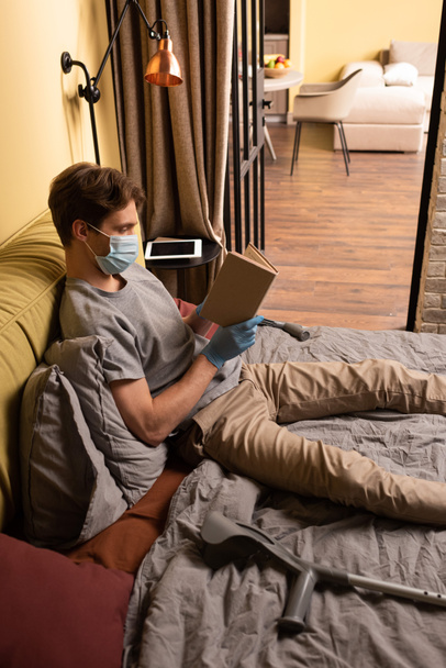 Homem com deficiência em máscara médica livro de leitura perto de muletas na cama
  - Foto, Imagem