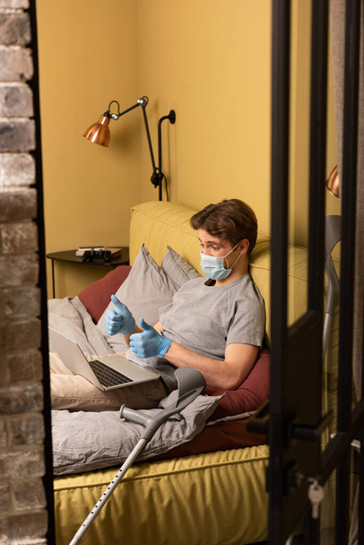 Vammaisen miehen valikoiva keskittyminen lääketieteelliseen naamioon, jossa näytetään peukalot videopuhelun aikana makuuhuoneessa kannettavalla tietokoneella
 - Valokuva, kuva