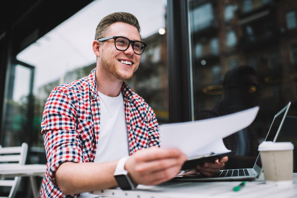 Happy young Caucasian man in eyeglasses holding clipboard with papers while sitting at table with laptop in outside cafe smiling away - 写真・画像
