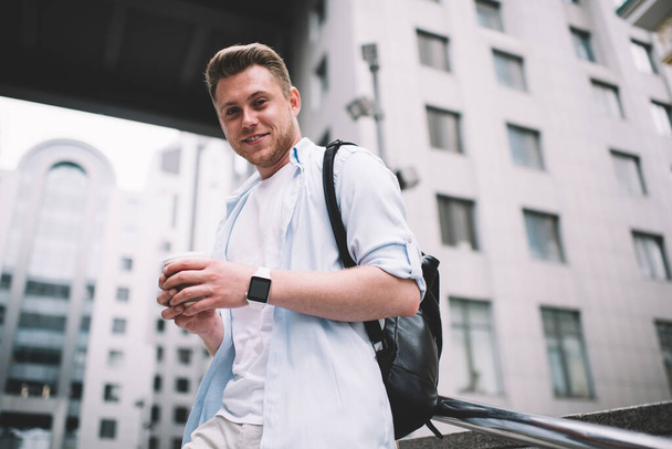 From below of handsome millennial man with bag holding paper cup of coffee and smiling at camera standing on street - Photo, image
