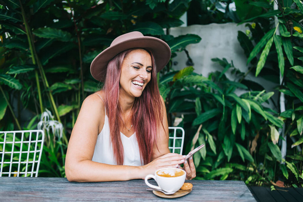 Cheerful mature tanned woman with dyed hair in hat using smartphone and laughing while sitting on table with cup of coffee - Photo, image