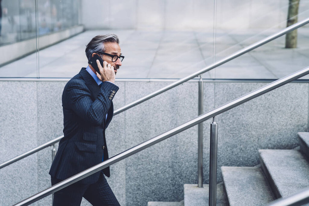 Side view of mature handsome grey-haired businessman in official suit and glasses calling and communicating while going up stairs at street in city - Foto, afbeelding