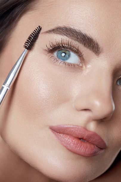 Makeup. Woman Brushing Eyebrows With Brush Close Up Portrait. Beautiful Brunette With Perfect Skin. Blue-Eyed Girl Looking Up.  - Valokuva, kuva