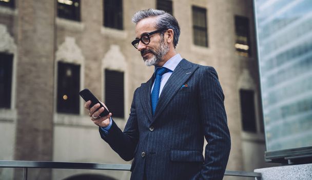 Middle aged bearded businessman in glasses and official wear standing on transparent balcony against grey skyscraper with smartphone in hand - Foto, imagen