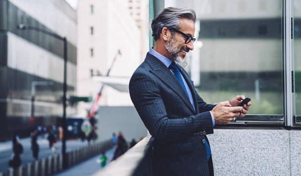Side view of happy middle aged man in official clothing and glasses standing on balcony and typing on mobile phone - 写真・画像