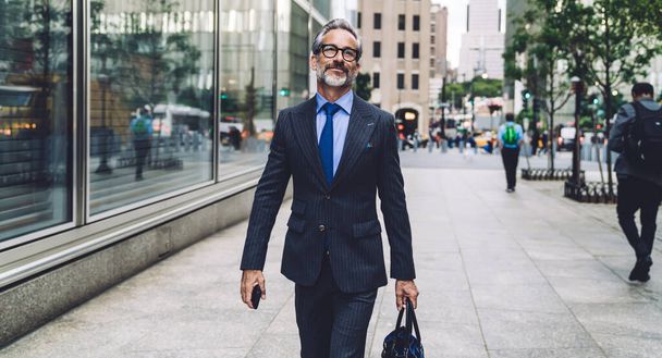 Successful content classy man in trendy suit and sunglasses holding smartphone and bag in hands while walking confidently on New York street - Fotografie, Obrázek