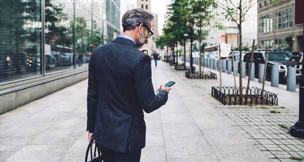 Back view of successful man in stylish dark striped business suit typing phone number in megapolis street in cloudy weather in New York - Fotó, kép