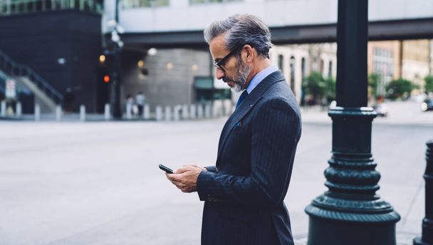 Side view of mature fashionable businessman tapping on smartphone standing on sidewalk by column in New York on blurred background - Photo, Image